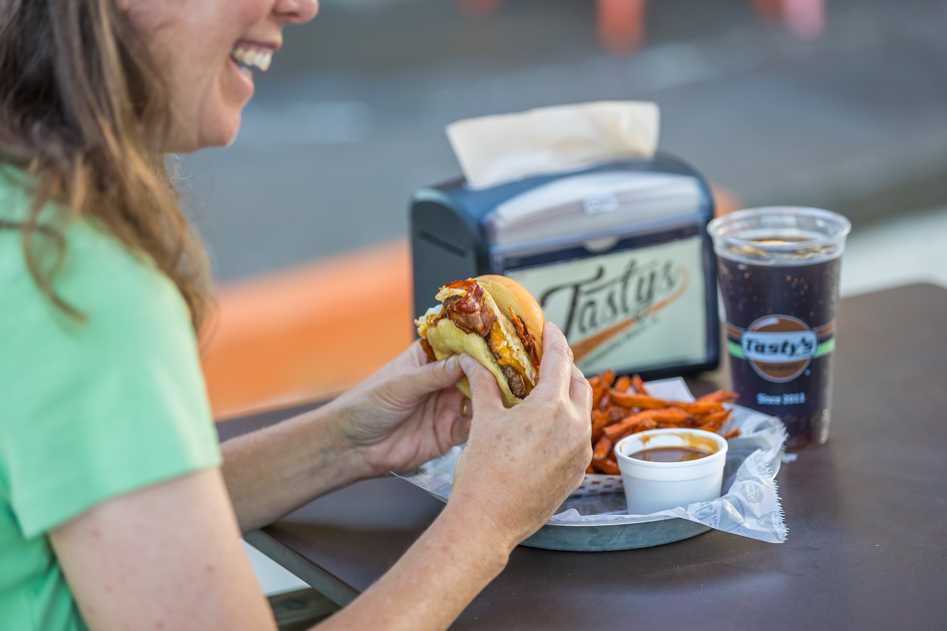 A woman in a green shirt smiles while holding a burger at an outdoor table, with a tray of sweet potato fries, a cup of sauce, a drink, and a napkin dispenser labeled Tastys in front of her.