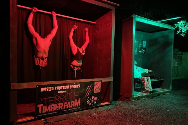 “A spooky Halloween display at the Terror on Timber Farm event in Nassau County features two lifelike dummies hanging upside down by their feet in a red-lit booth labeled ‘Terror on the Timber Farm,’ with a figure dressed in white seated in a nearby green-lit booth.