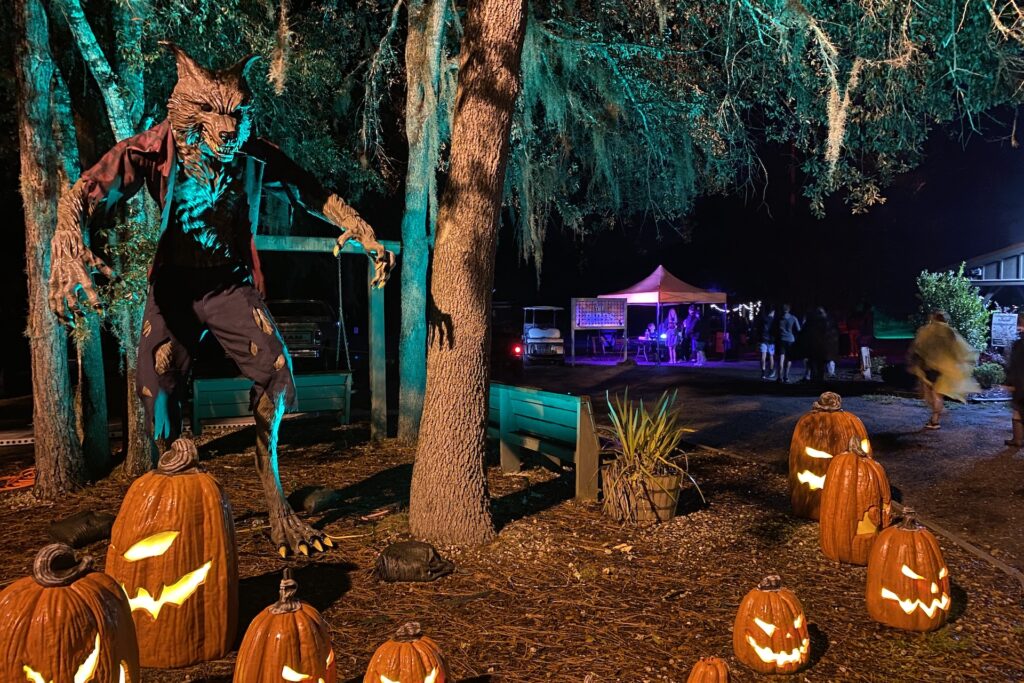 A large werewolf figure stands among glowing jack-o-lanterns under trees at night. In the background, people gather near a tent lit with purple lights, creating a spooky Halloween scene.