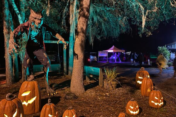 A large werewolf figure stands among glowing jack-o’-lanterns under moss-covered trees at night during the Terror on Timber Farm Halloween event in Nassau County. In the background, people gather near a tent lit with purple lights, creating a spooky festival atmosphere.