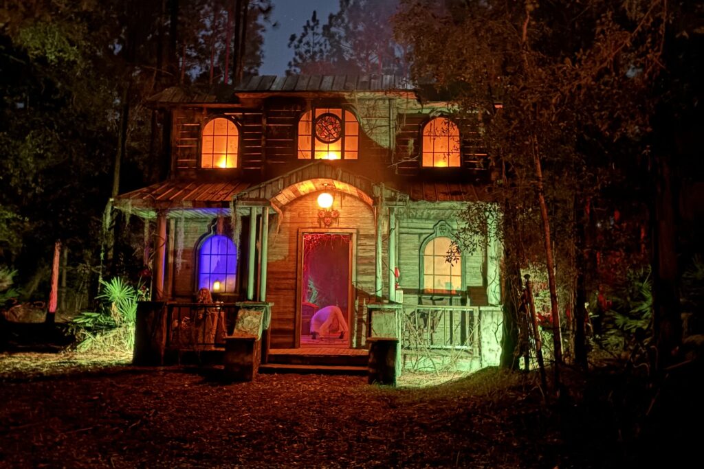 A two-story wooden house in the woods at night during the Terror on Timber Farm Halloween event in Nassau County, illuminated by eerie orange, green, and purple lights. The glowing windows and rustic porch create a spooky, haunted atmosphere.