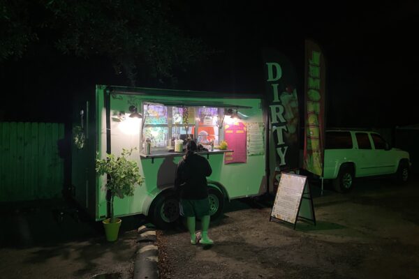 A person stands at a brightly lit food truck at night during the Terror on Timber Farm Halloween event in Nassau County. The truck glows green with a menu, a small potted plant, and flags reading ‘DIRTY’ and ‘LEMONADE,’ with a white SUV parked nearby.
