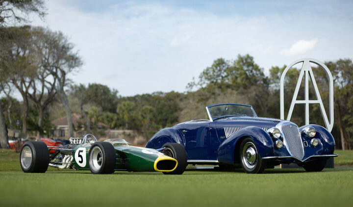 A green and yellow vintage race car with the number 5 is parked next to a blue classic convertible on a grassy field, with trees and a large sculpture in the background.