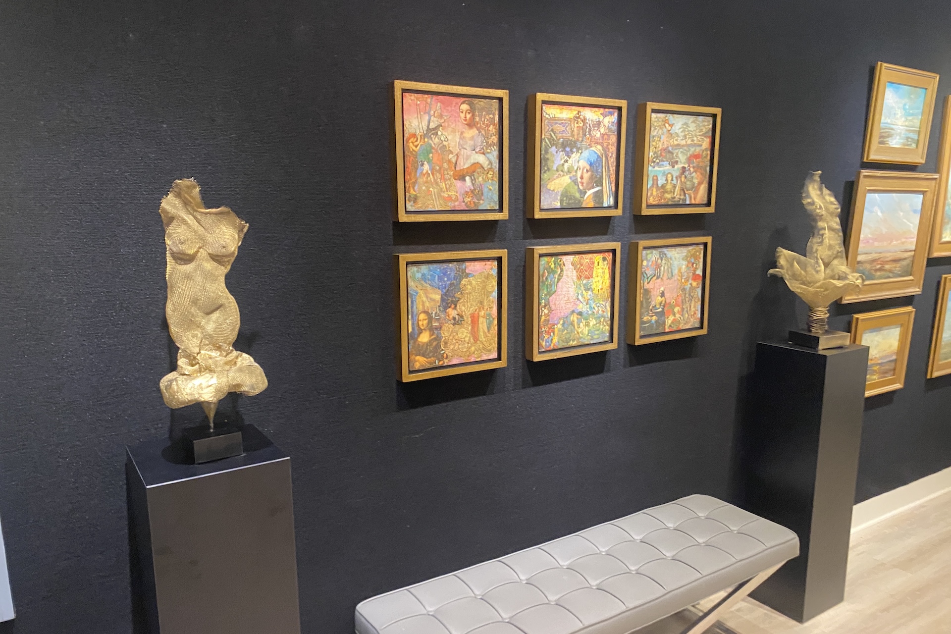 Golden sculptures on black pedestals and a bench are in front of a dark wall displaying colorful paintings arranged in a grid. Several framed landscape artworks are also visible on the wall.