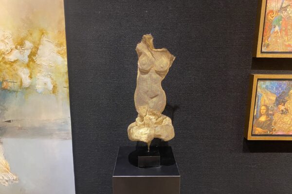 A textured golden sculpture of a female torso is displayed on a black pedestal against a dark wall, with abstract and colorful framed paintings hanging on either side.