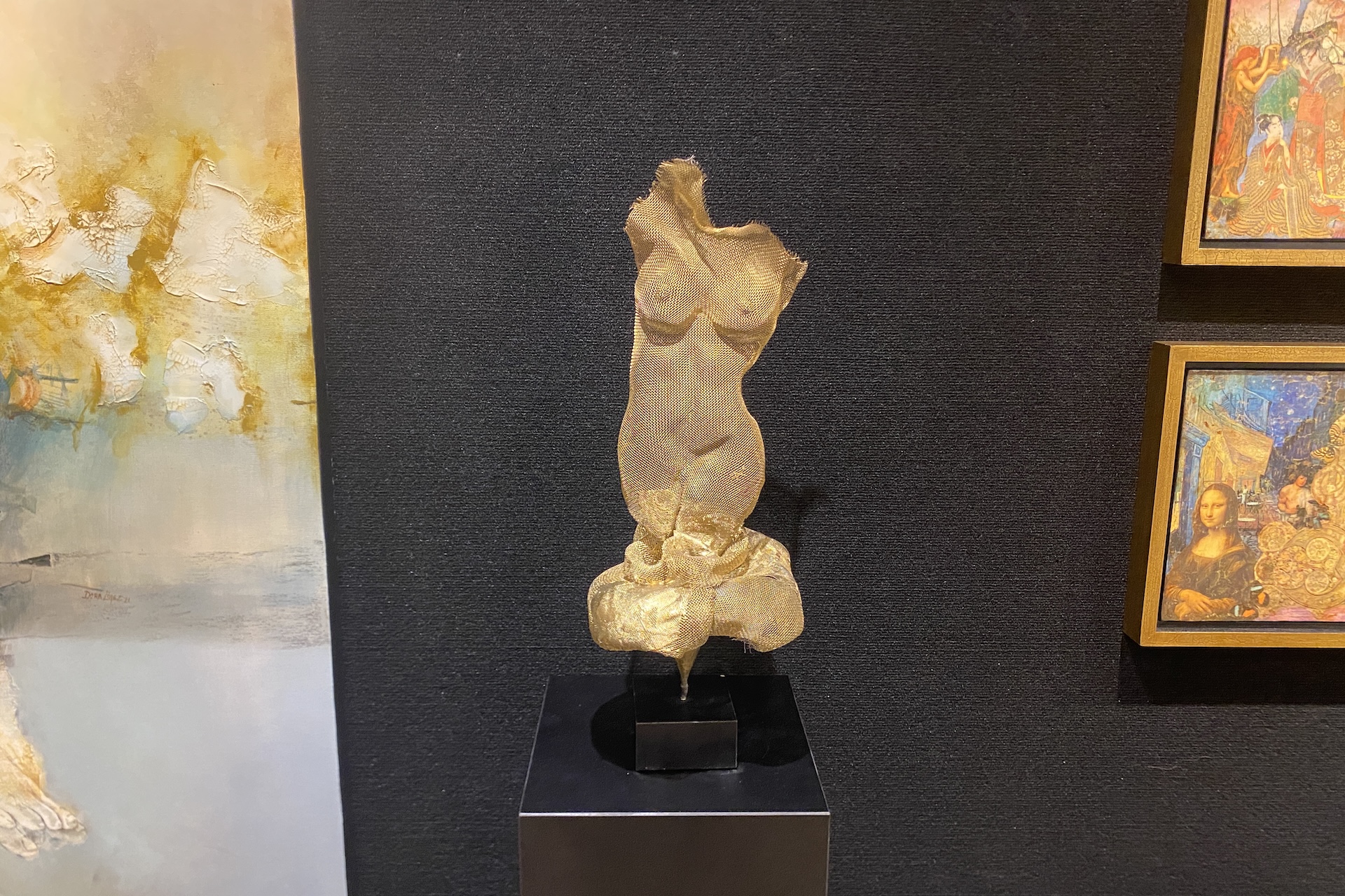 A textured golden sculpture of a female torso is displayed on a black pedestal against a dark wall, with abstract and colorful framed paintings hanging on either side.