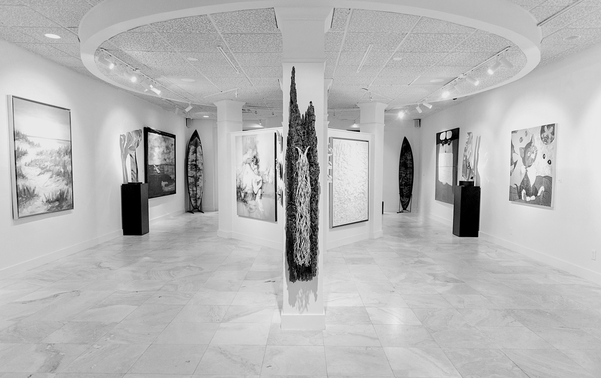 A bright, modern art gallery displays various paintings and sculptures on white walls and pedestals, with artwork arranged around a central pillar and along the curved room.
