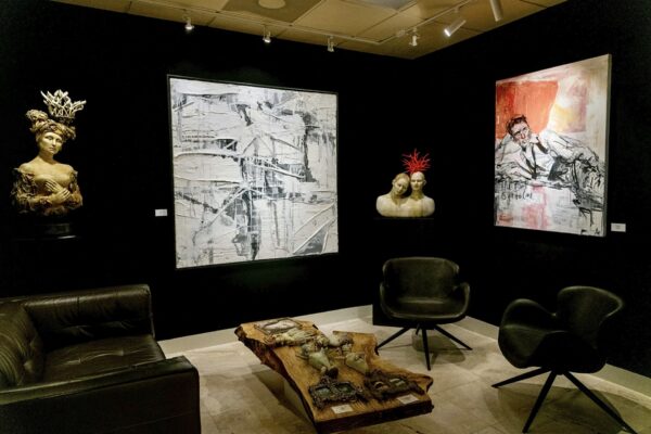 A modern art gallery room with two abstract paintings on black walls, two sculpted busts with decorative headpieces, black leather chairs, and a wooden coffee table displaying art objects.