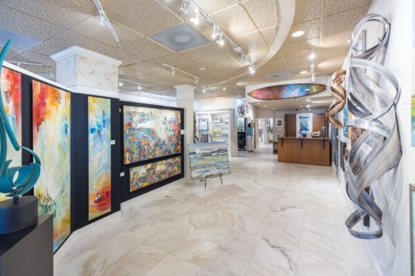 Bright art gallery with colorful abstract paintings on the walls, contemporary sculptures, and a tiled floor. A reception desk is visible in the back, and the space is well-lit with ceiling lights.