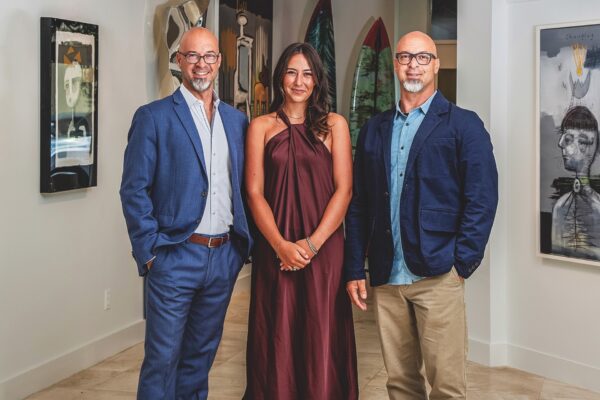 Three people pose indoors in front of artwork: two men in blue suits and glasses stand on either side of a woman in a sleeveless maroon dress, all smiling. Paintings and surfboards are visible in the background.