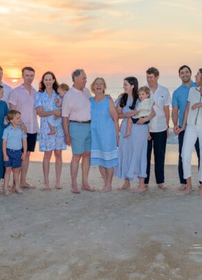 A multigenerational family, dressed in pastel and blue clothing, stands barefoot on a sandy beach at sunset. They smile and interact with each other, with gentle waves and a colorful sky in the background.
