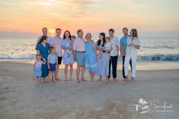 A multigenerational family, dressed in pastel and blue clothing, stands barefoot on a sandy beach at sunset. They smile and interact with each other, with gentle waves and a colorful sky in the background.