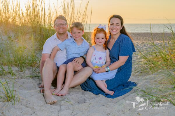 A family of four sits smiling on sandy beach grass at sunset, with the ocean in the background. The parents are seated on either side of their young son and daughter, who wear light summer clothes.