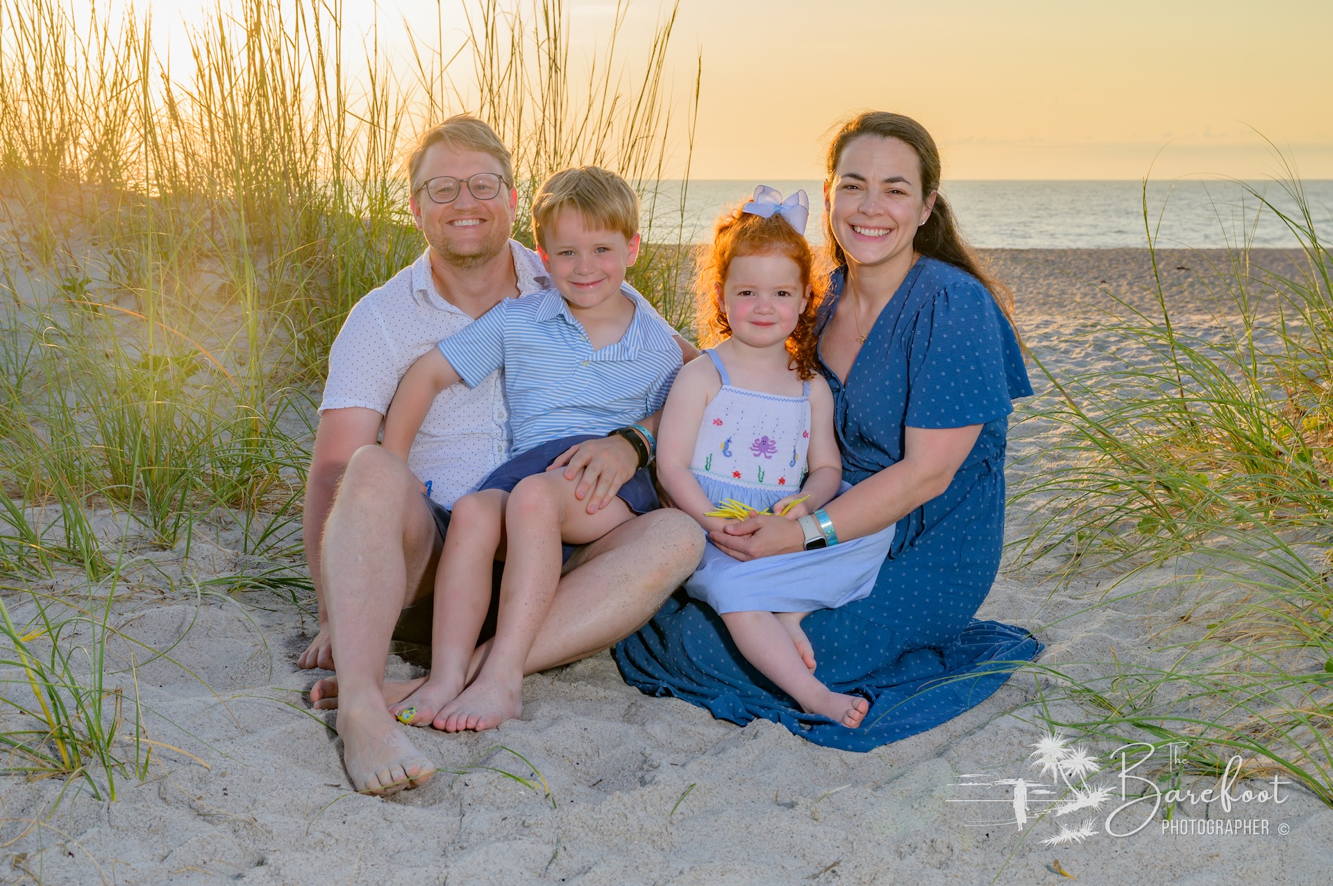 A family of four sits smiling on sandy beach grass at sunset, with the ocean in the background. The parents are seated on either side of their young son and daughter, who wear light summer clothes.