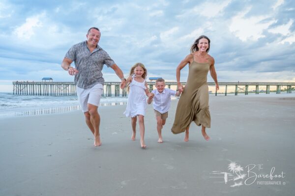 A smiling family of four—two adults and two children—run barefoot along a sandy beach near the shoreline, with a pier and cloudy sky in the background. Everyone appears joyful and relaxed.