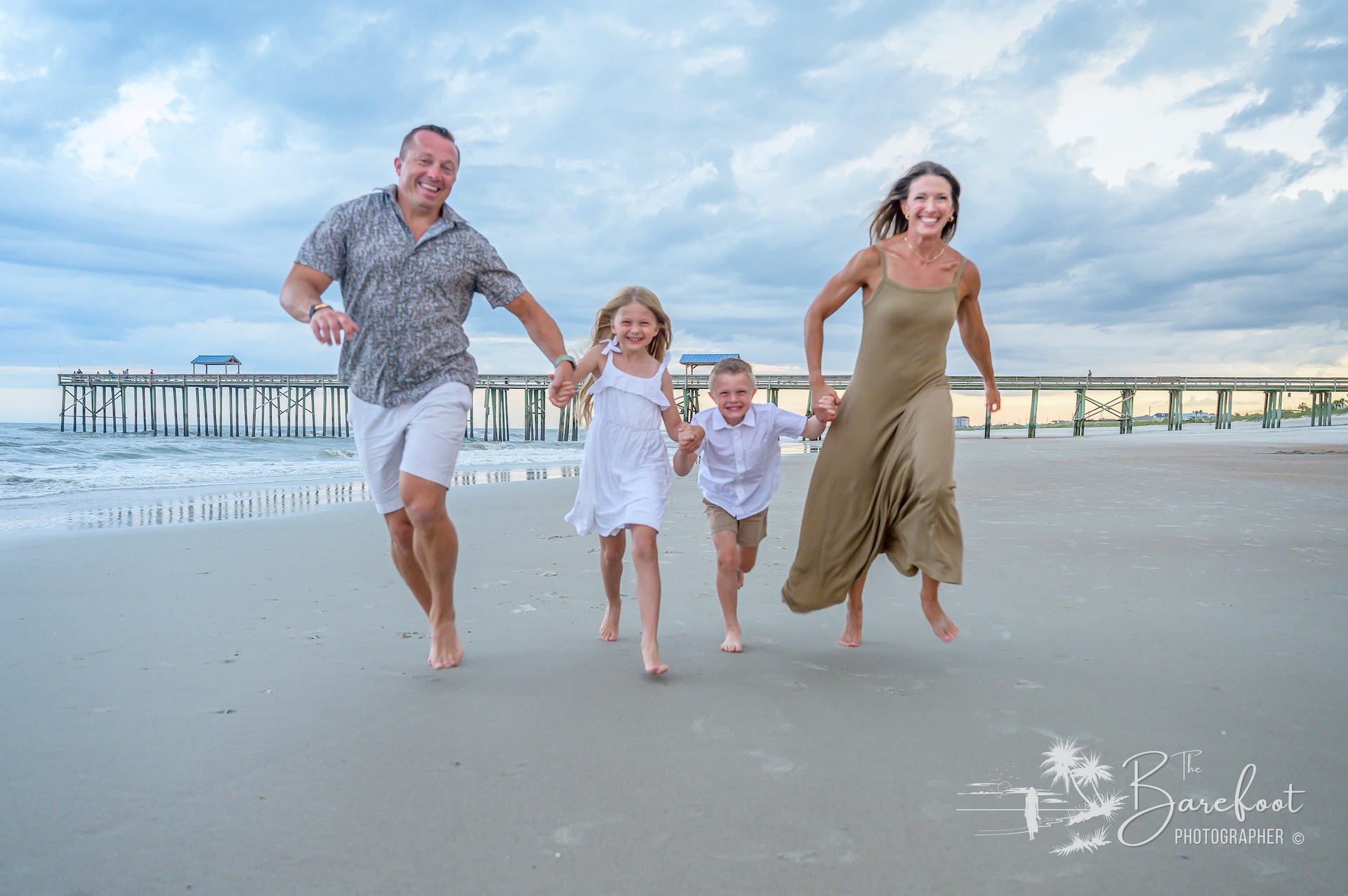 A smiling family of four—two adults and two children—run barefoot along a sandy beach near the shoreline, with a pier and cloudy sky in the background. Everyone appears joyful and relaxed.