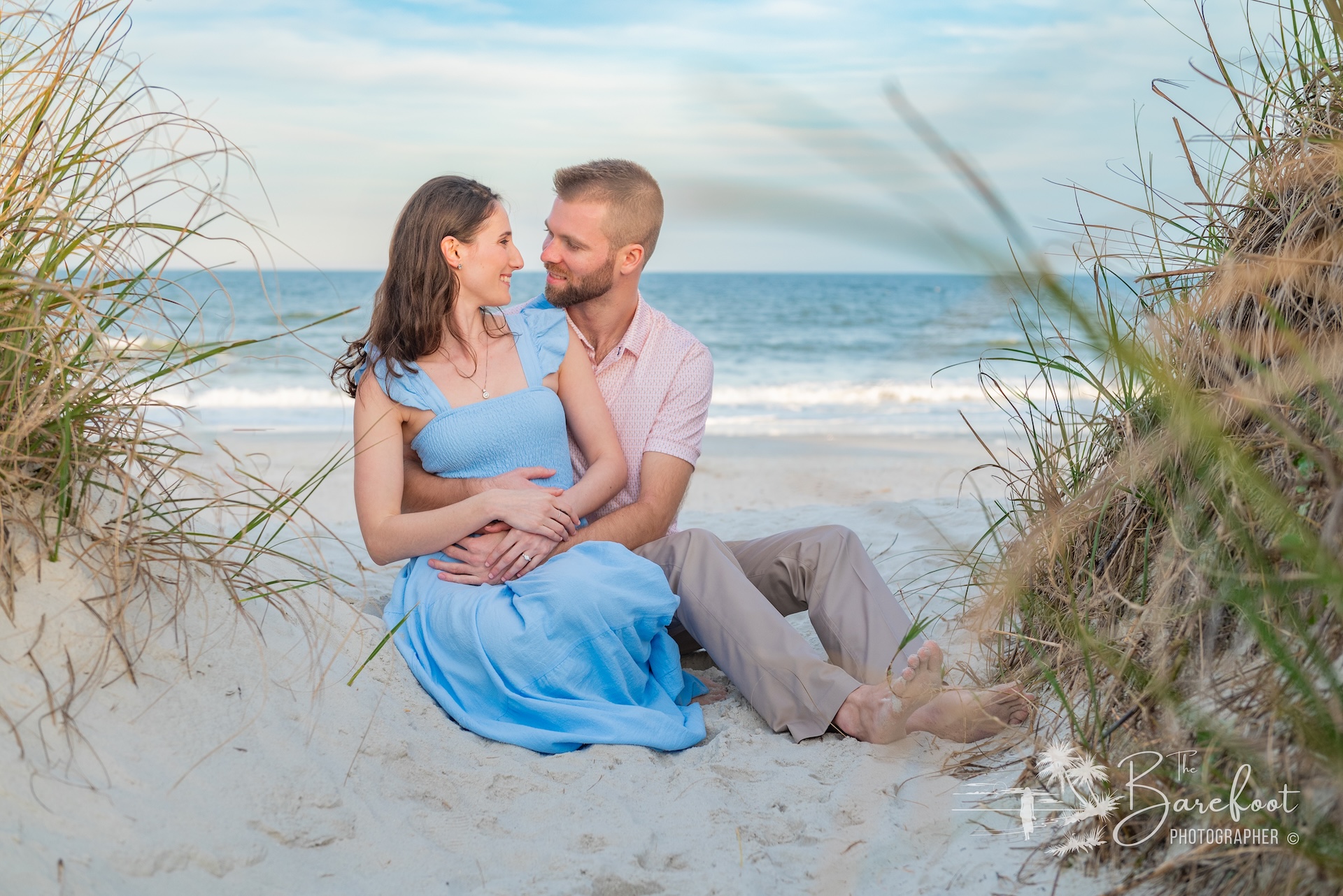A couple sits together on sandy beach surrounded by beach grass, smiling and gazing at each other with the ocean in the background. The woman wears a light blue dress, and the man a pale shirt and tan pants.