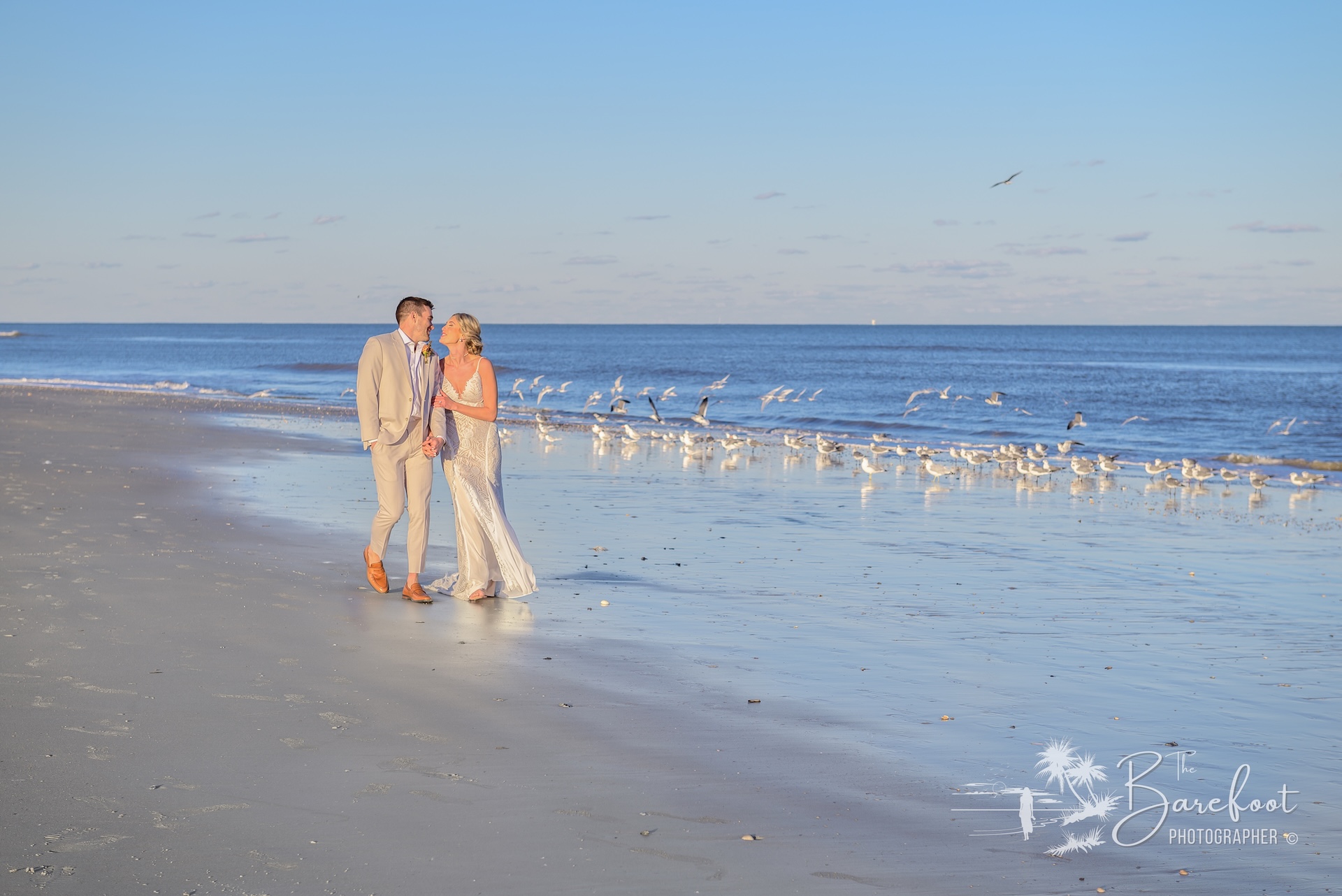 A couple dressed in formal wedding attire walks hand in hand along a sandy beach, with gentle waves and a flock of seabirds in the background under a clear blue sky.