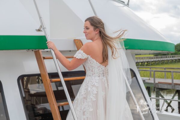 A bride in an off-the-shoulder lace wedding dress and veil stands on a boat, holding a wooden ladder, with greenery visible in the background.