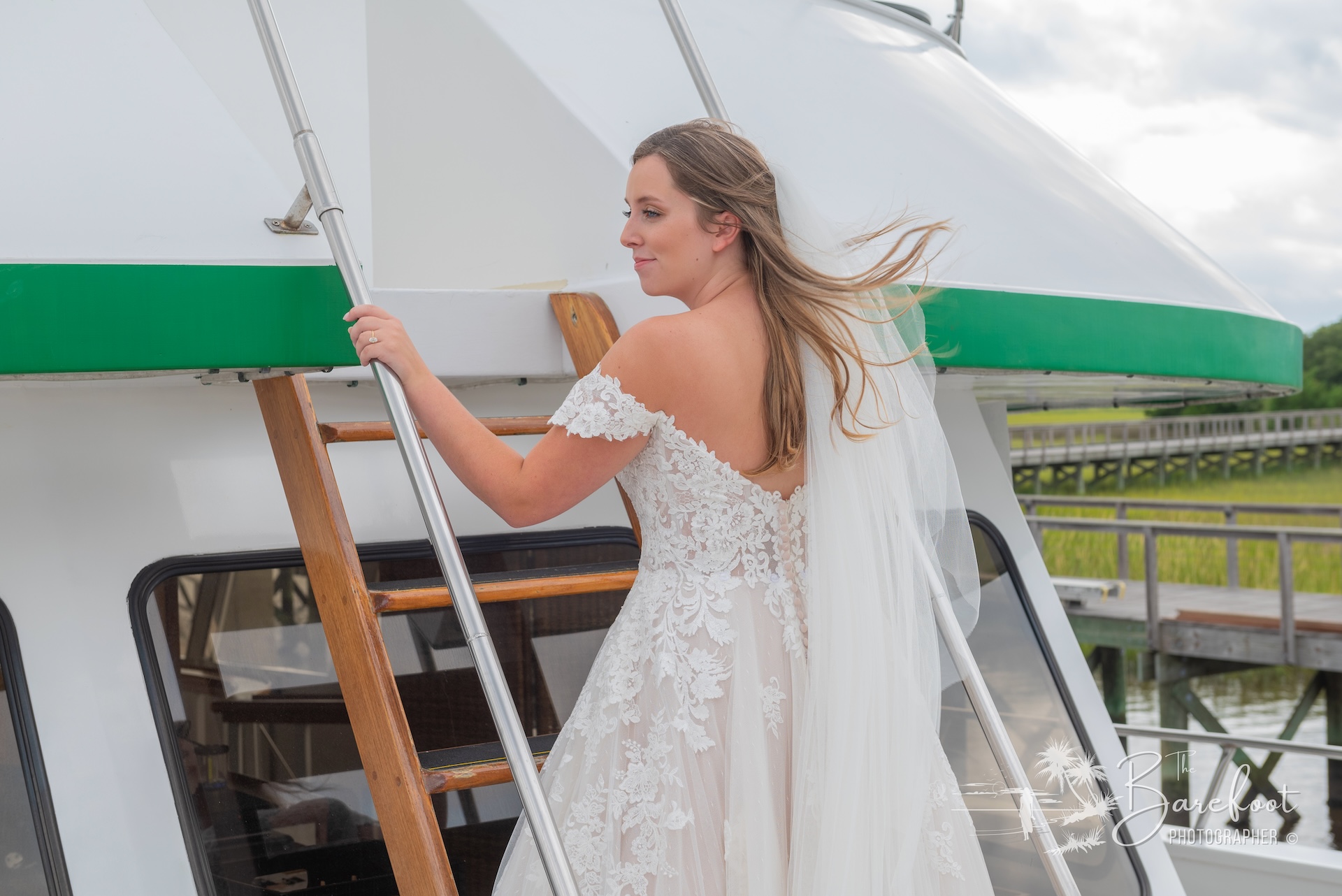 A bride in an off-the-shoulder lace wedding dress and veil stands on a boat, holding a wooden ladder, with greenery visible in the background.