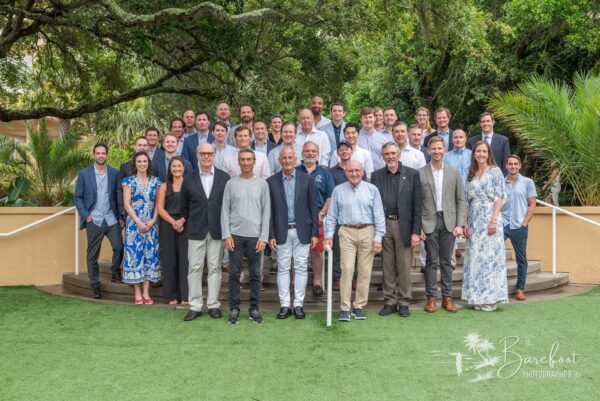 A large group of men and women, dressed in business casual and formal attire, pose for a group photo outdoors on green grass with trees and greenery in the background.