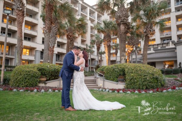 A bride and groom kiss on a grassy lawn in front of a resort building lined with palm trees. The bride wears a white gown, and the groom is in a navy suit. Flowers and greenery surround them.