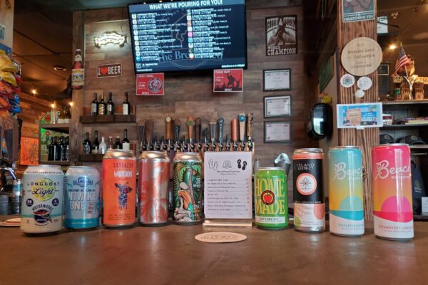 A row of colorful beer cans is displayed on a bar counter with taps and various decorations behind them, and a digital beer list is visible on a screen above.