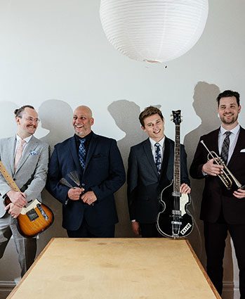 Four men in suits stand indoors, smiling, each holding a musical instrument: an electric guitar, drumsticks, a bass guitar, and a trumpet. A round white hanging lamp is above them. A wooden table is in the foreground.