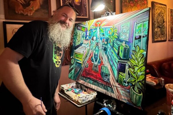 A man with a long gray beard smiles while painting a vibrant, colorful room scene on a canvas in an art-filled studio. His palette is filled with mixed paints.