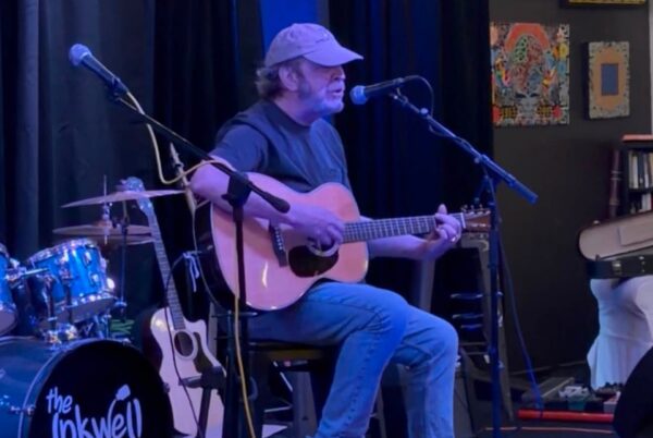 A man wearing a cap and casual clothes plays an acoustic guitar and sings into a microphone on stage. Drums and other guitars are visible, along with colorful artwork and books in the background.