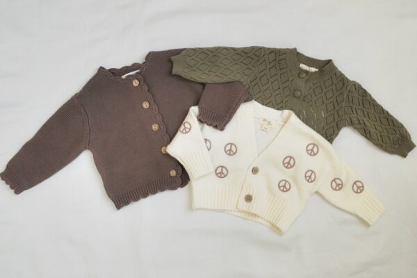 Three baby cardigans are laid out on a white surface: one brown with scalloped edges and wooden buttons, one olive green with a diamond pattern, and one cream with peace signs and tan buttons.