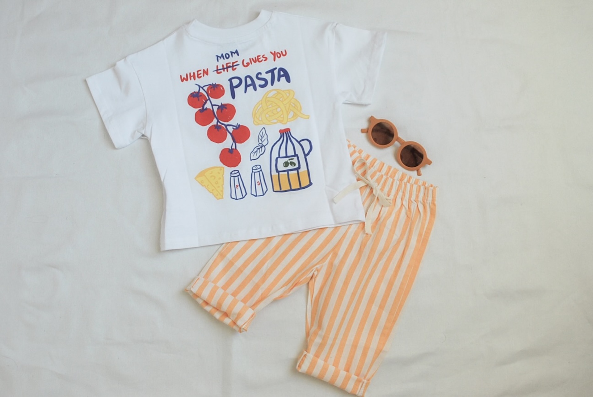A childrens outfit with a white T-shirt featuring food drawings and the text Mom, when life gives you pasta, paired with orange-and-white striped pants and brown sunglasses, laid out on a white background.