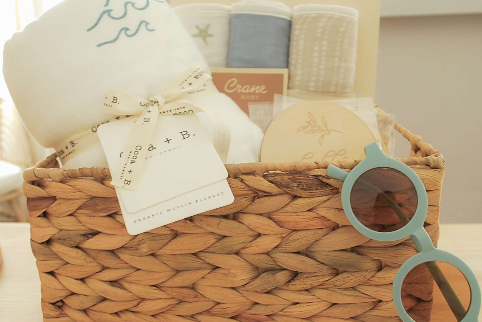 A woven basket filled with baby items including blankets, a round wooden brush, and light blue baby sunglasses. One blanket is tied with ribbon and has a tag that reads a + b organic muslin blankets.