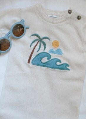 A white baby romper with embroidered designs of a palm tree, wave, sun, and clouds, laid on a white cushion next to a pair of round blue sunglasses.