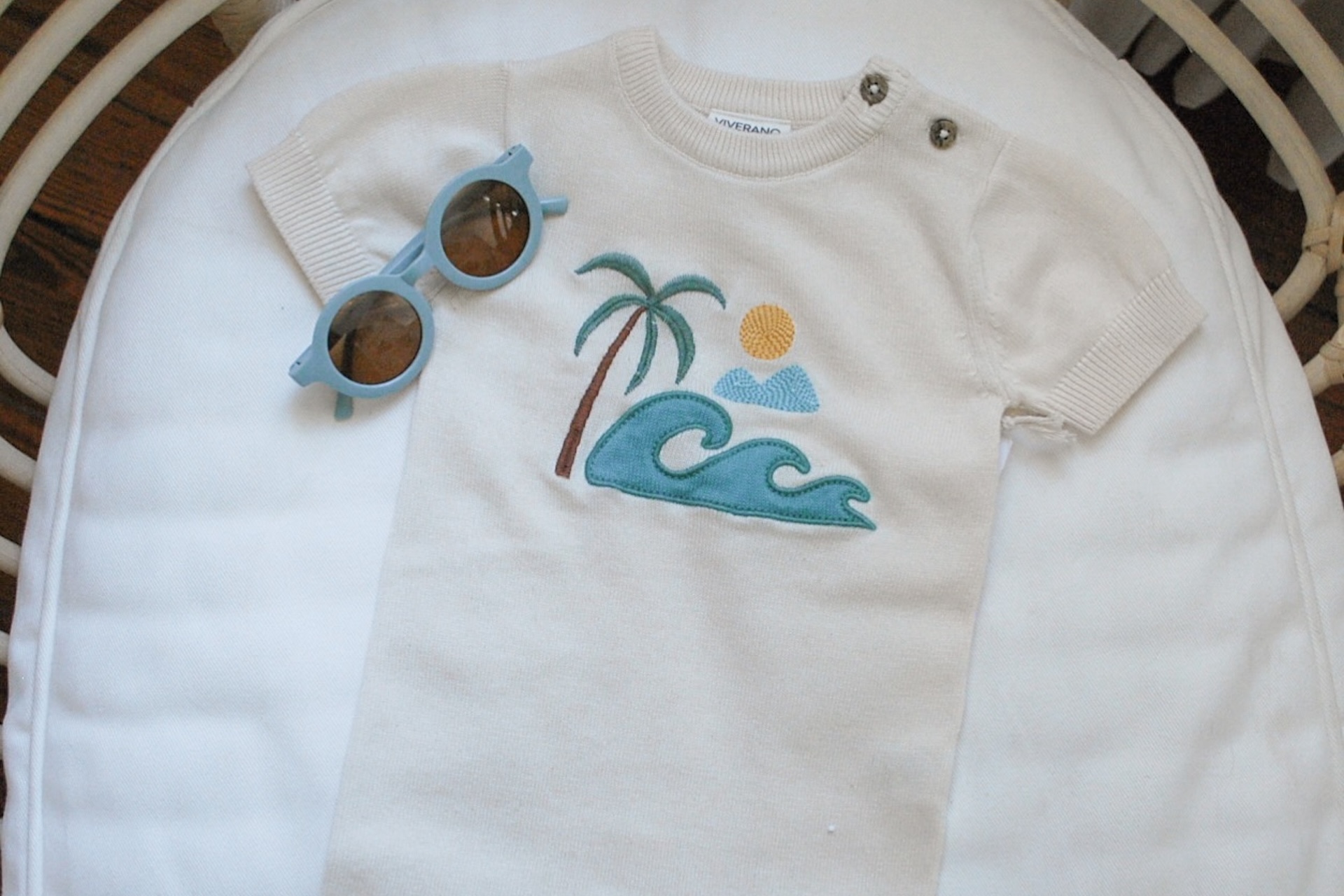 A white baby romper with embroidered designs of a palm tree, wave, sun, and clouds, laid on a white cushion next to a pair of round blue sunglasses.