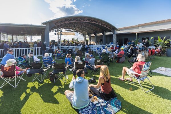 A large group of people sit on lawn chairs and blankets on artificial grass, watching a performance under an outdoor pavilion on a sunny day. Some eat food and socialize, enjoying the relaxed atmosphere.