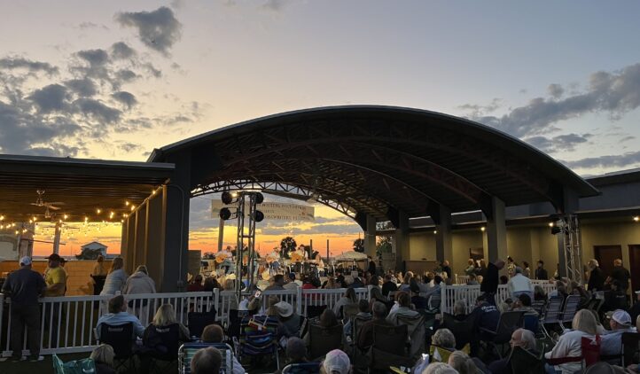 A crowd sits on lawn chairs and blankets, watching a live outdoor concert at sunset under a large, curved pavilion. String lights hang nearby, and the sky is filled with clouds and orange hues.