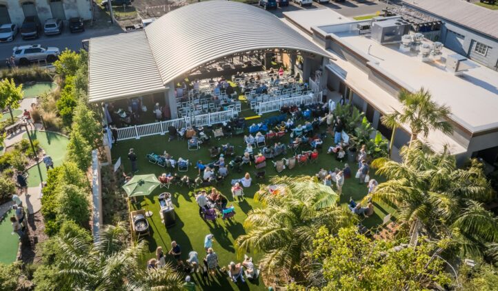 Aerial view of an outdoor event with people sitting on grass and chairs, gathered around a stage with a curved roof. Surrounding areas have palm trees, buildings, and parked cars. The atmosphere is lively and sunny.