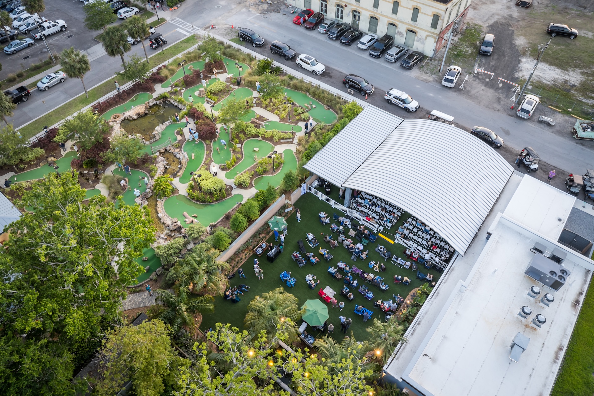 Aerial view of a mini golf course with winding green paths next to an outdoor event space filled with people seated under a large white canopy, surrounded by trees and parked cars.