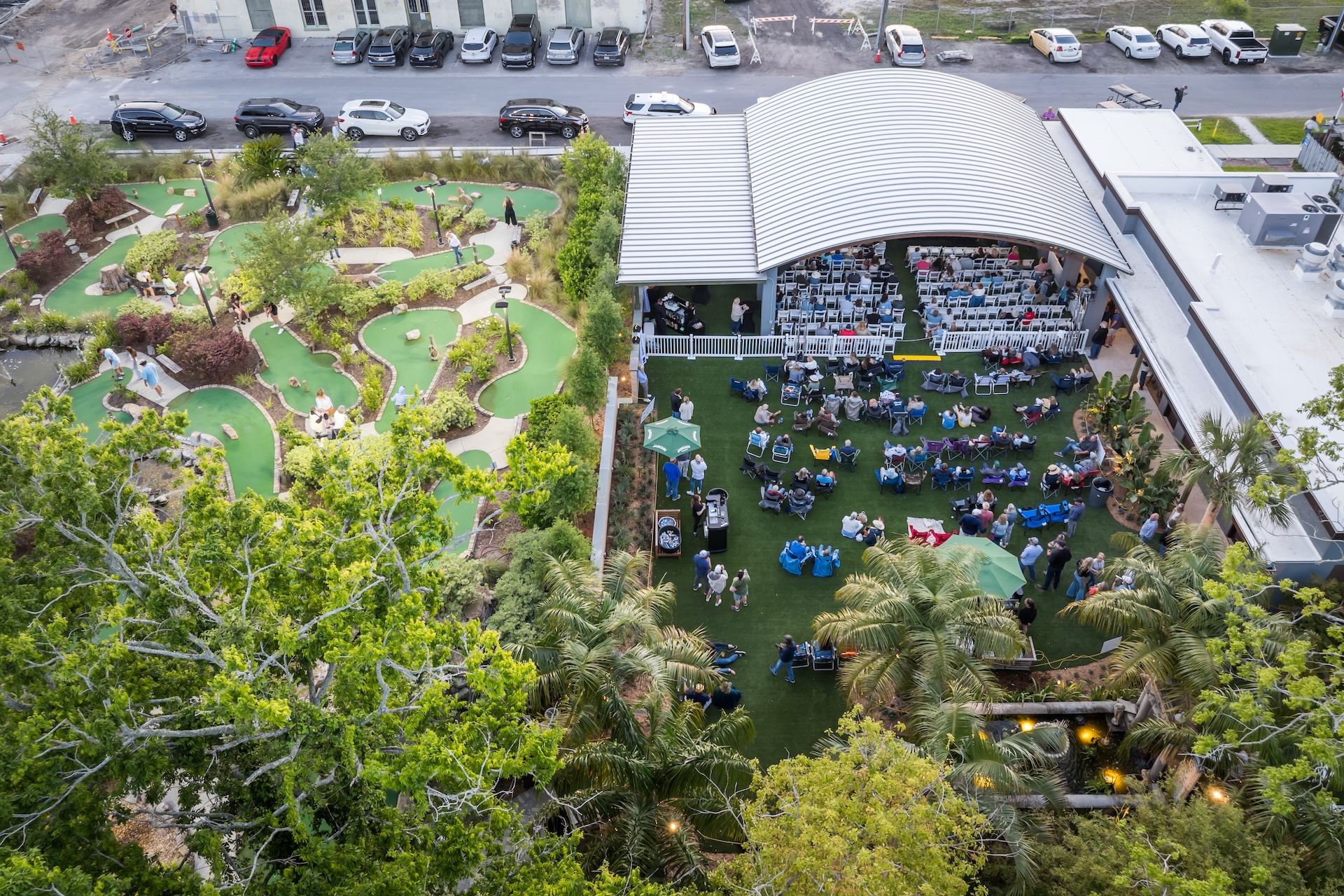 Aerial view of a venue with a mini-golf course on the left and an outdoor event space on the right, where people are seated in rows under a large, curved roof. Trees surround the area, and parked cars line the street.