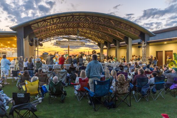 A crowd watches a live outdoor concert under a large, open-air pavilion at sunset. People sit on lawn chairs or stand, facing the stage where musicians perform. The sky is partly cloudy, creating a vibrant and lively atmosphere.