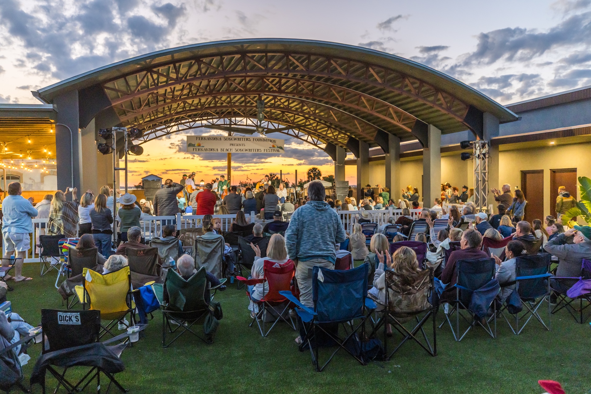 A crowd watches a live outdoor concert under a large, open-air pavilion at sunset. People sit on lawn chairs or stand, facing the stage where musicians perform. The sky is partly cloudy, creating a vibrant and lively atmosphere.