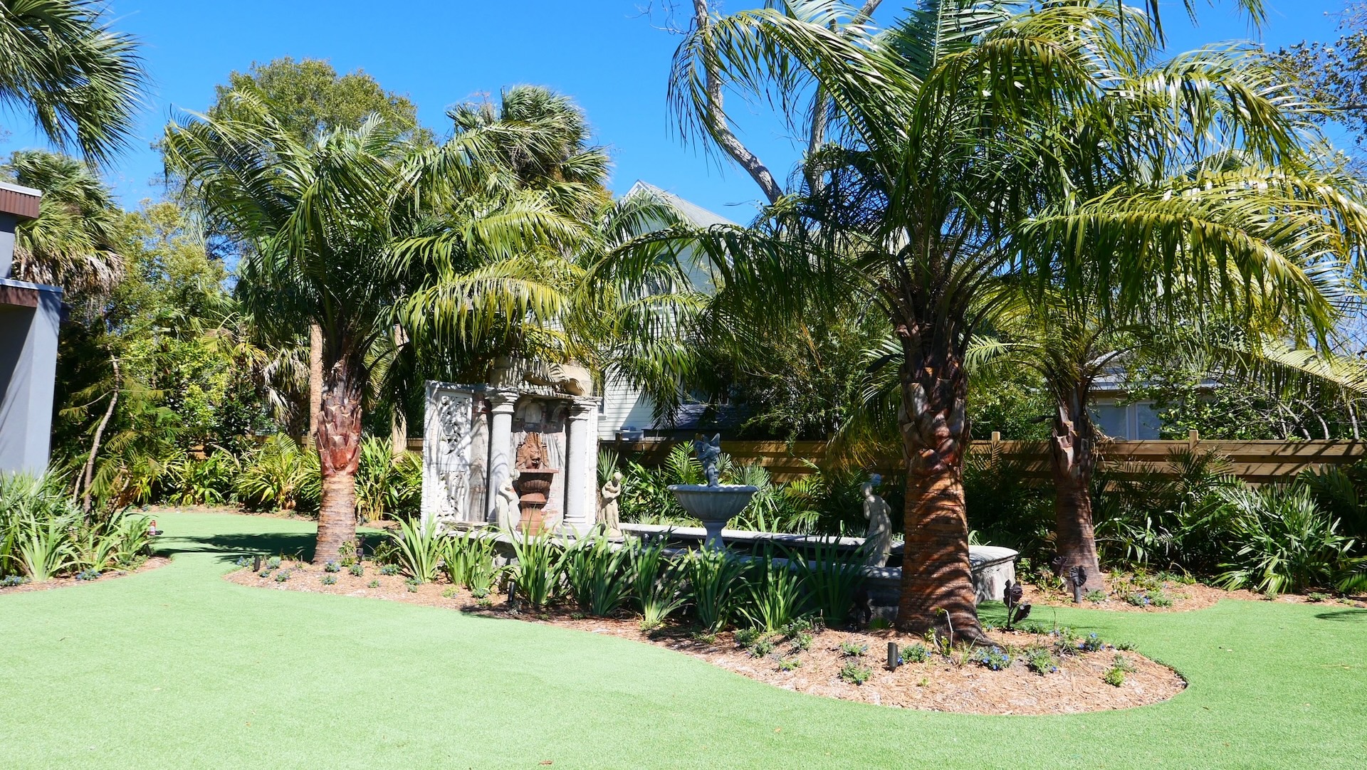 A lush garden with palm trees, manicured grass, and a stone fountain centerpiece surrounded by plants, set under a clear blue sky.