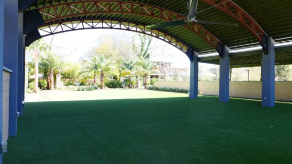 A covered outdoor area with a green artificial turf floor, blue pillars, and an arched metal roof. Sunlight and trees are visible beyond the open side.