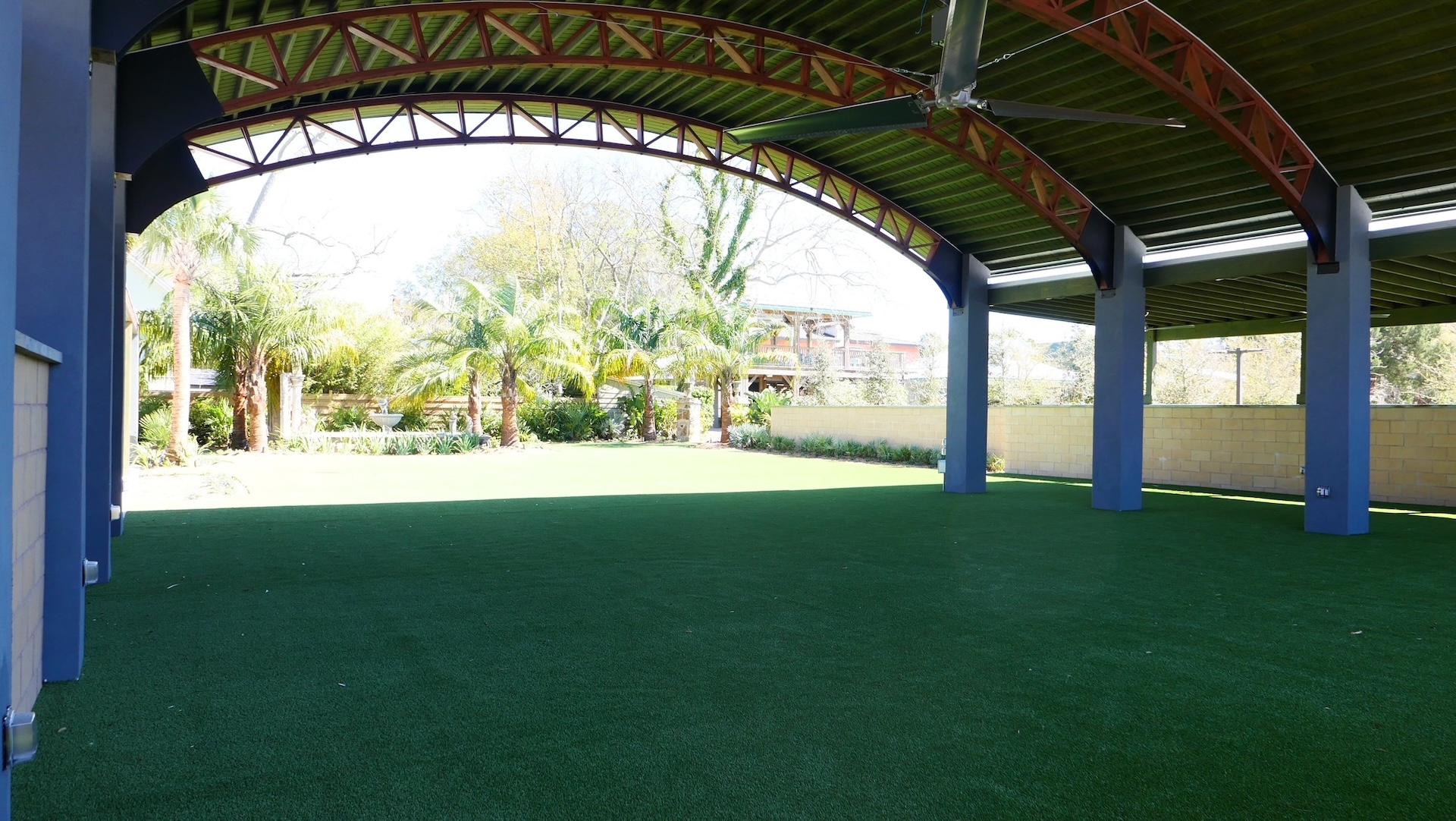 A covered outdoor area with a green artificial turf floor, blue pillars, and an arched metal roof. Sunlight and trees are visible beyond the open side.