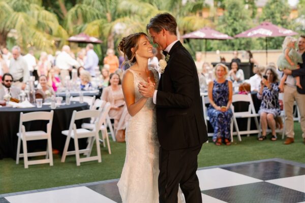 A bride and groom share their first dance at an outdoor wedding reception, smiling at each other. Guests sit and watch from white chairs, with palm trees and umbrellas in the background.