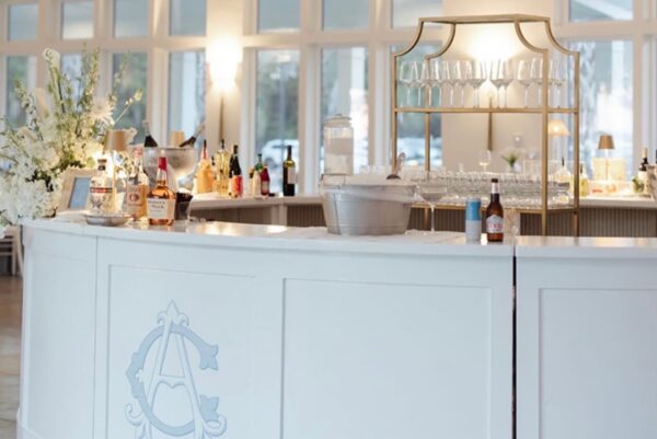 A white curved bar with glassware, bottles of liquor, ice bucket, and floral arrangements. Soft natural light streams in through large windows in the background.