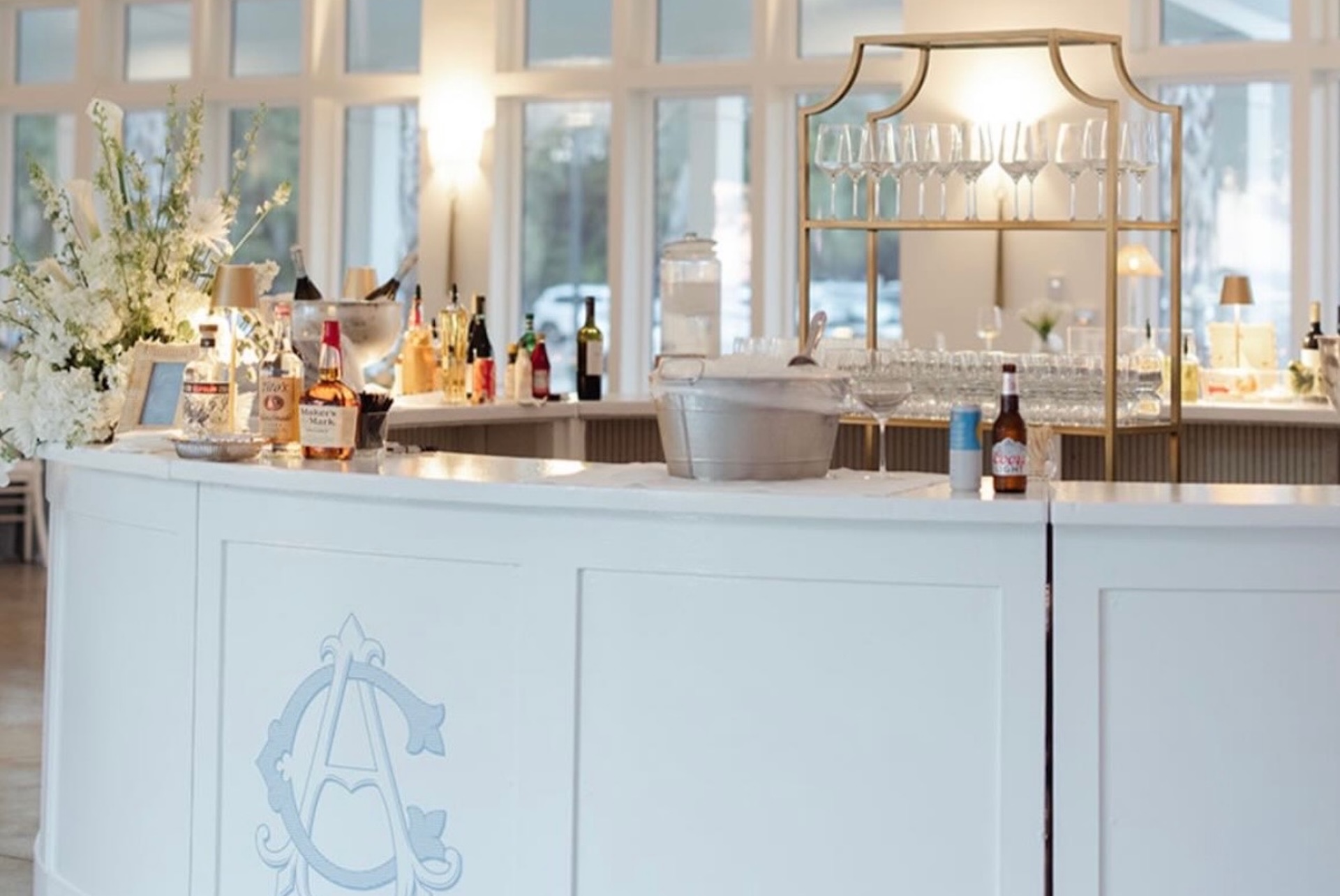 A white curved bar with glassware, bottles of liquor, ice bucket, and floral arrangements. Soft natural light streams in through large windows in the background.