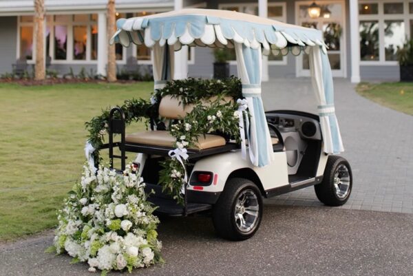 A white golf cart decorated with draped fabric, green garlands, and white flowers is parked on a path in front of a building with large windows and a lawn.