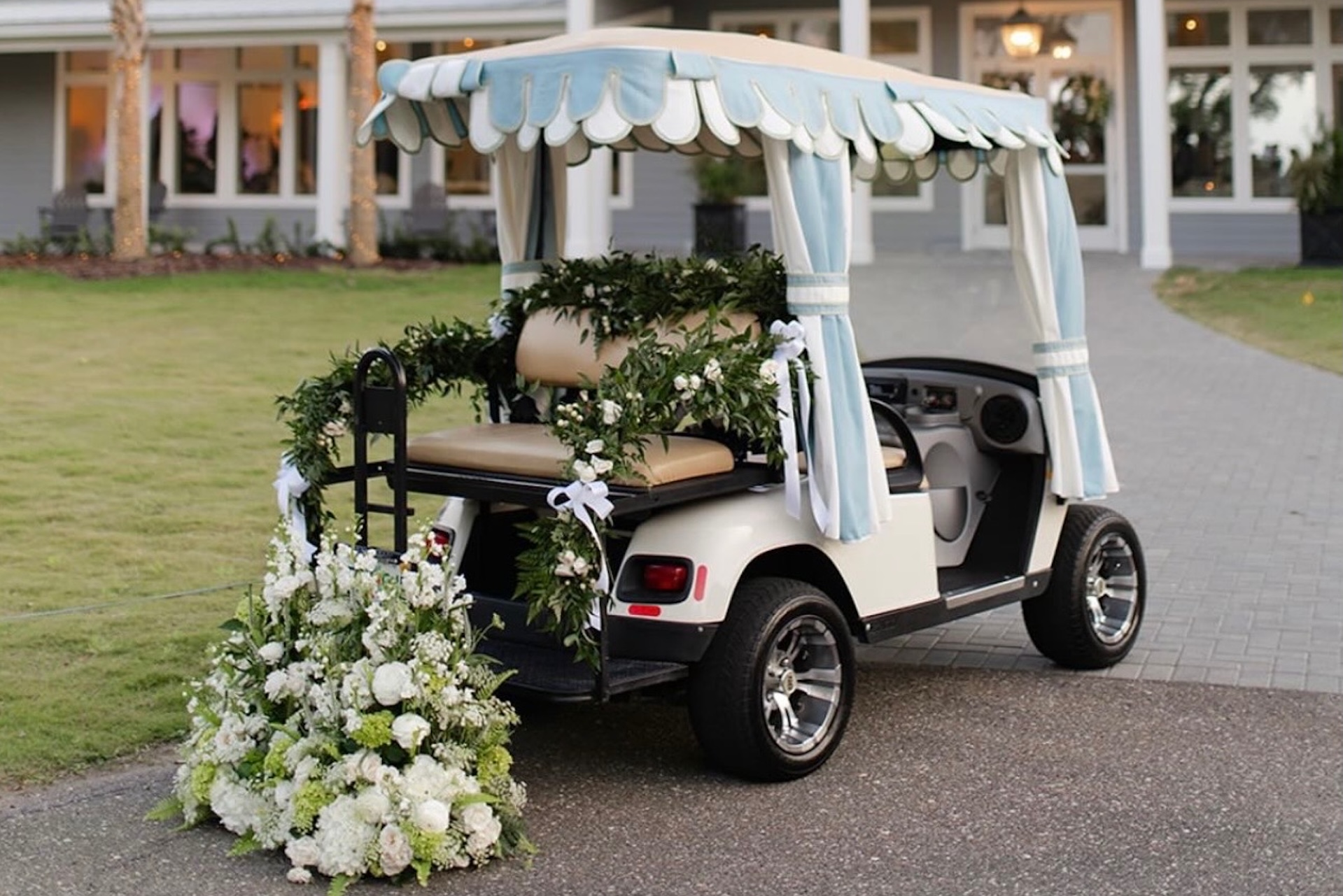 A white golf cart decorated with draped fabric, green garlands, and white flowers is parked on a path in front of a building with large windows and a lawn.